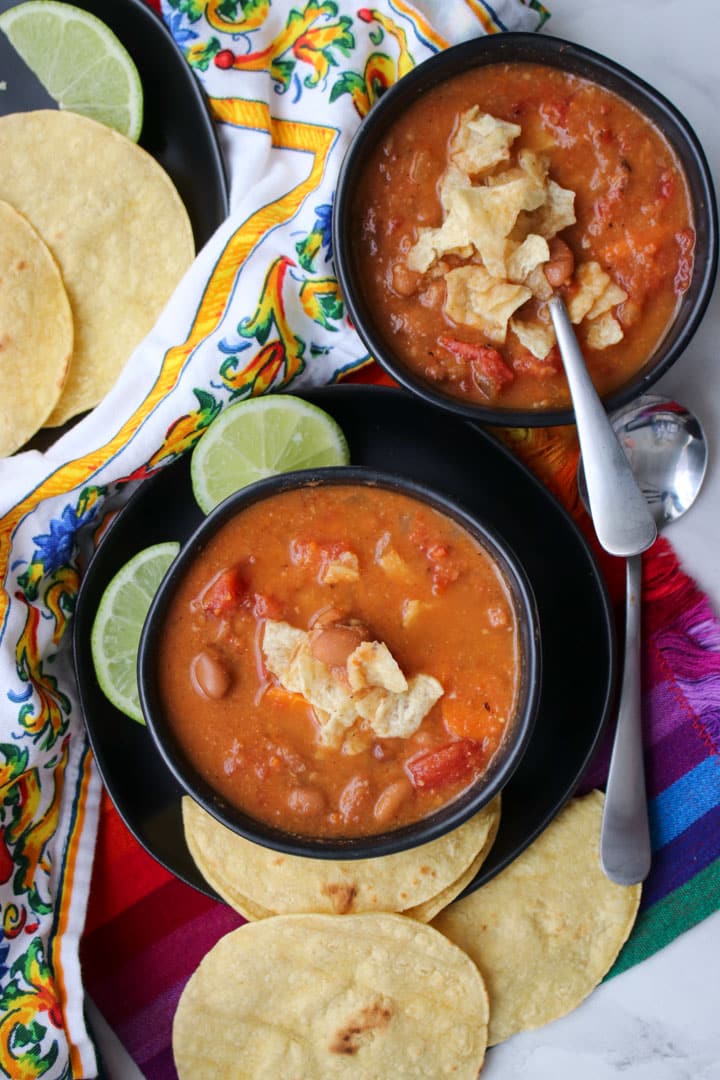 2 black bowls filled with pinto bean soup topped with tortilla chips and garnished with lime slices