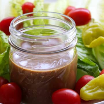 jar of brown creamy balsamic dressing surrounded by lettuce, tomatoes, peppers