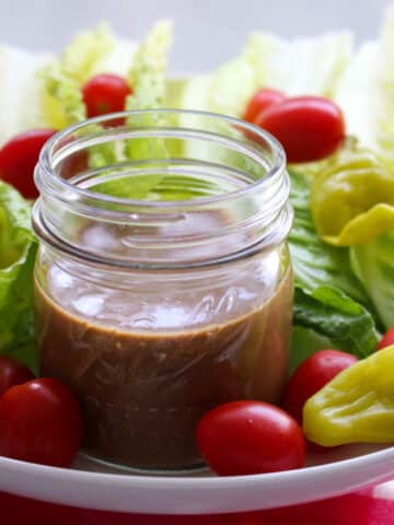 jar of brown creamy balsamic dressing surrounded by lettuce, tomatoes, peppers