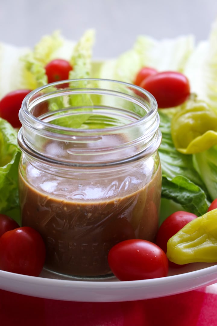 jar of brown creamy balsamic dressing surrounded by lettuce, tomatoes, peppers