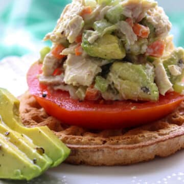 avocado chicken salad on top of a waffle and tomato slice with extra avocado on the side