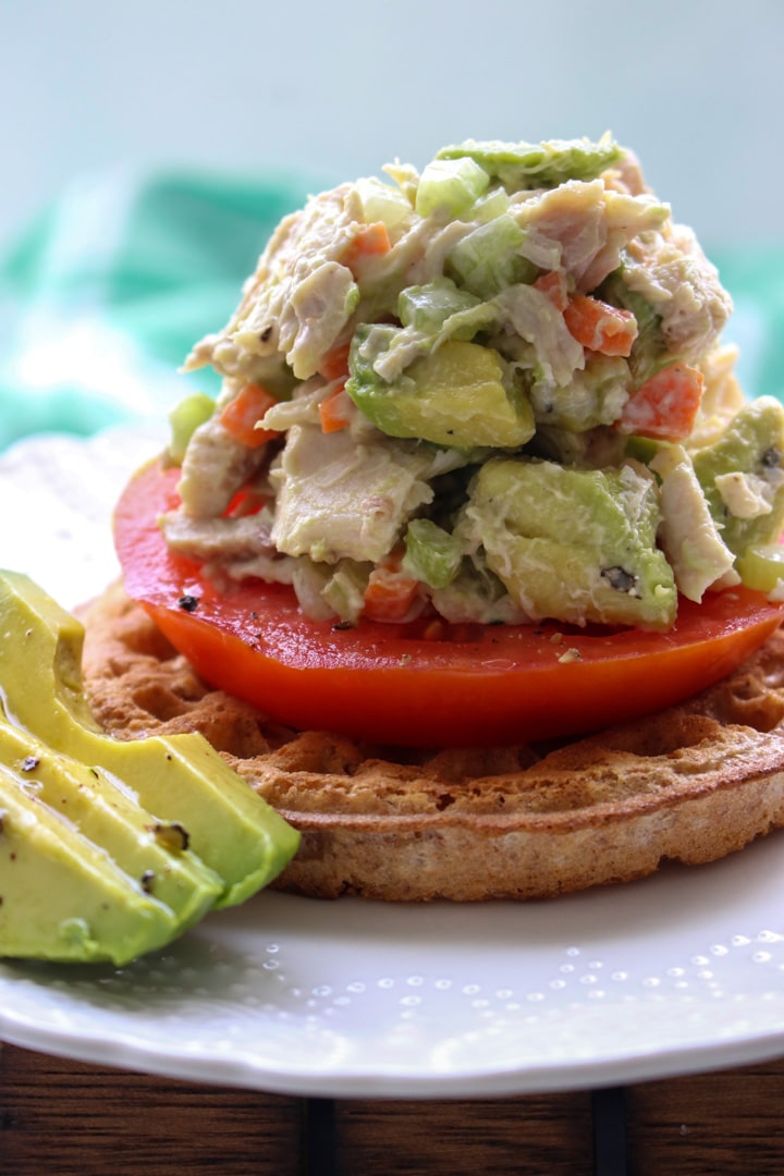 avocado chicken salad on top of a waffle and tomato slice with extra avocado on the side