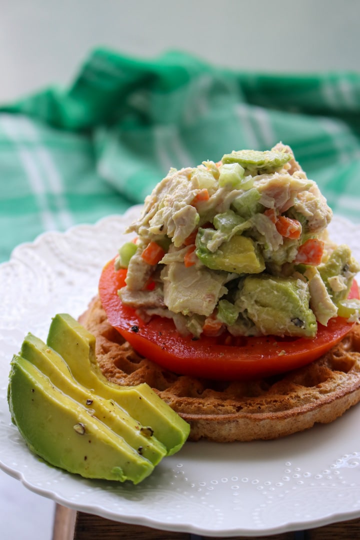 avocado chicken salad on top of a waffle and tomato slice with extra avocado on the side